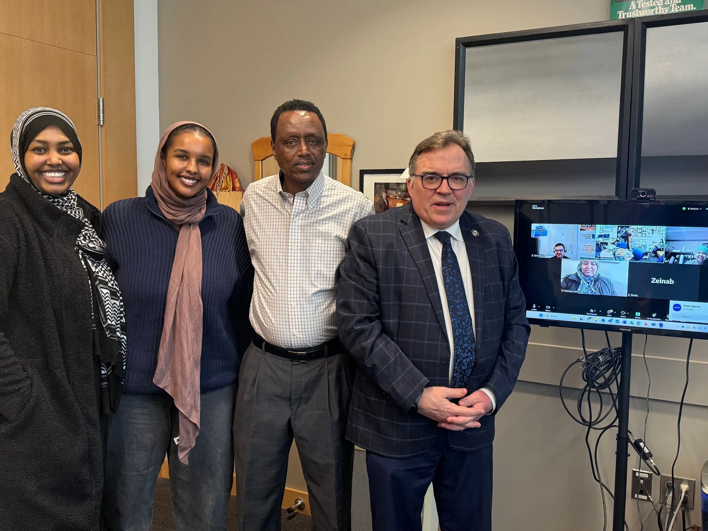 Four Maangaar Voices members stand with a man in a navy suit beside a video call screen.