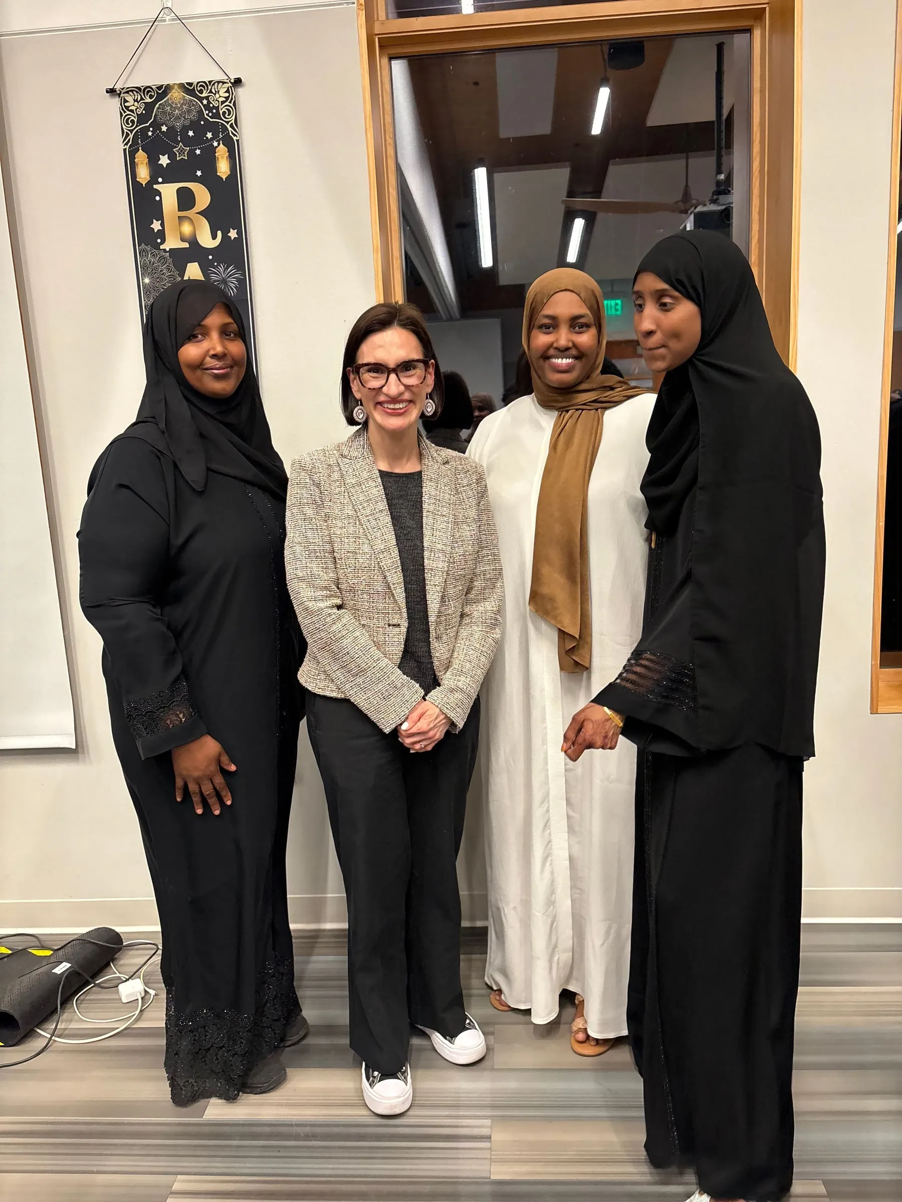 Three Maangaar Voices members stand with a woman in a tweed blazer beneath a Ramadan banner.