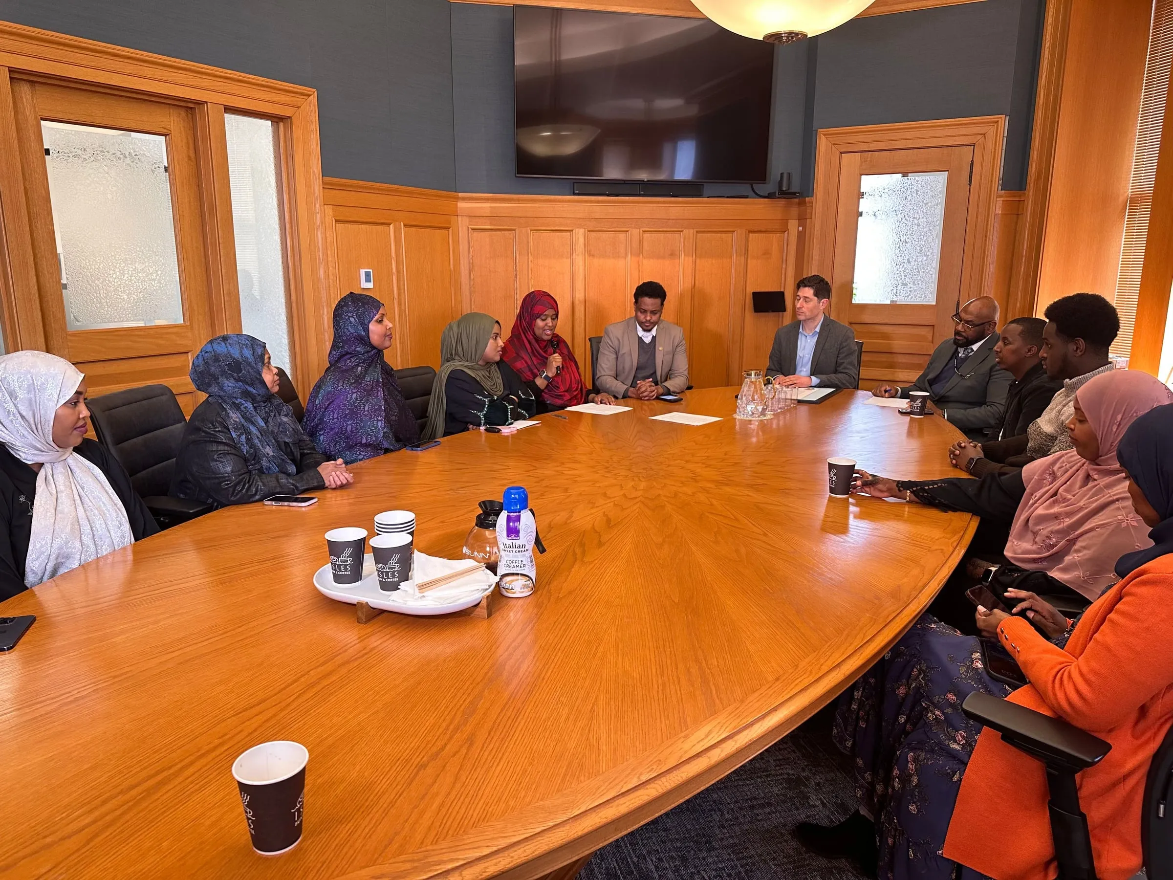 Maangaar Voices members and families sit around a long wooden conference table while a woman in a red hijab speaks into a microphone.