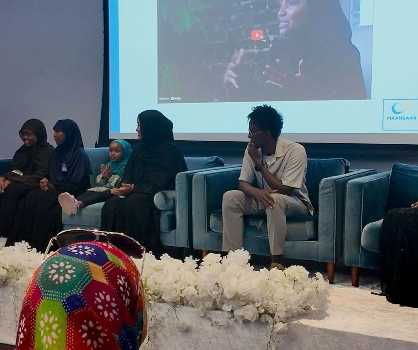 Sibling Panel members on blue velvet couches, with a young child among them, as a video plays on the screen behind the stage.