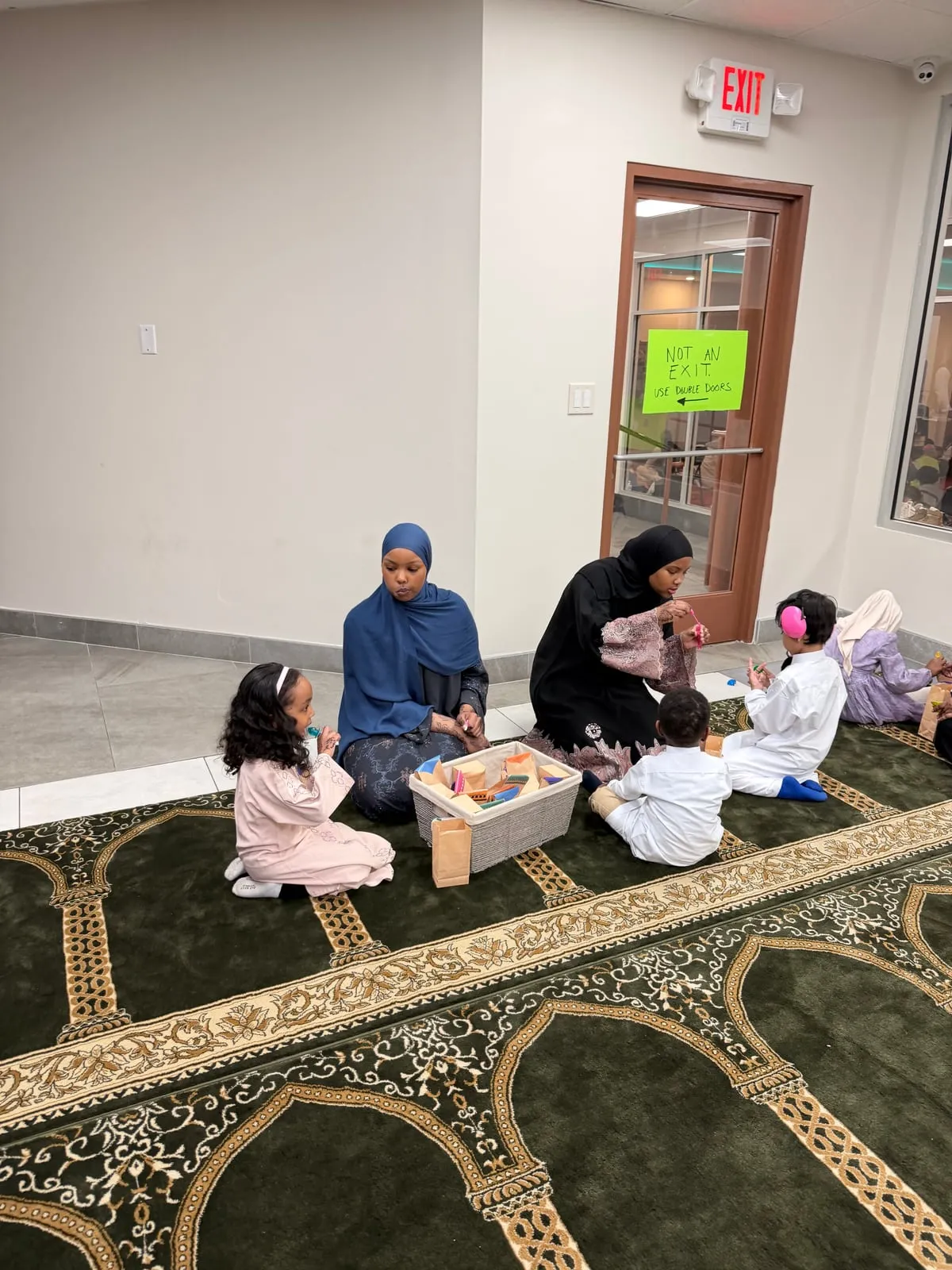 Several families sit together on the green masjid carpet while children open their sensory bags.