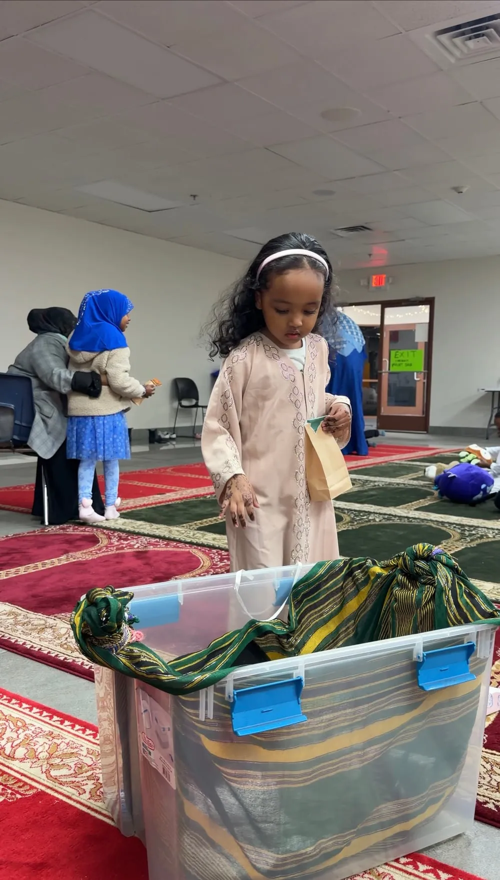 A girl in a cream dress reaches into a sensory bin on the prayer rug.