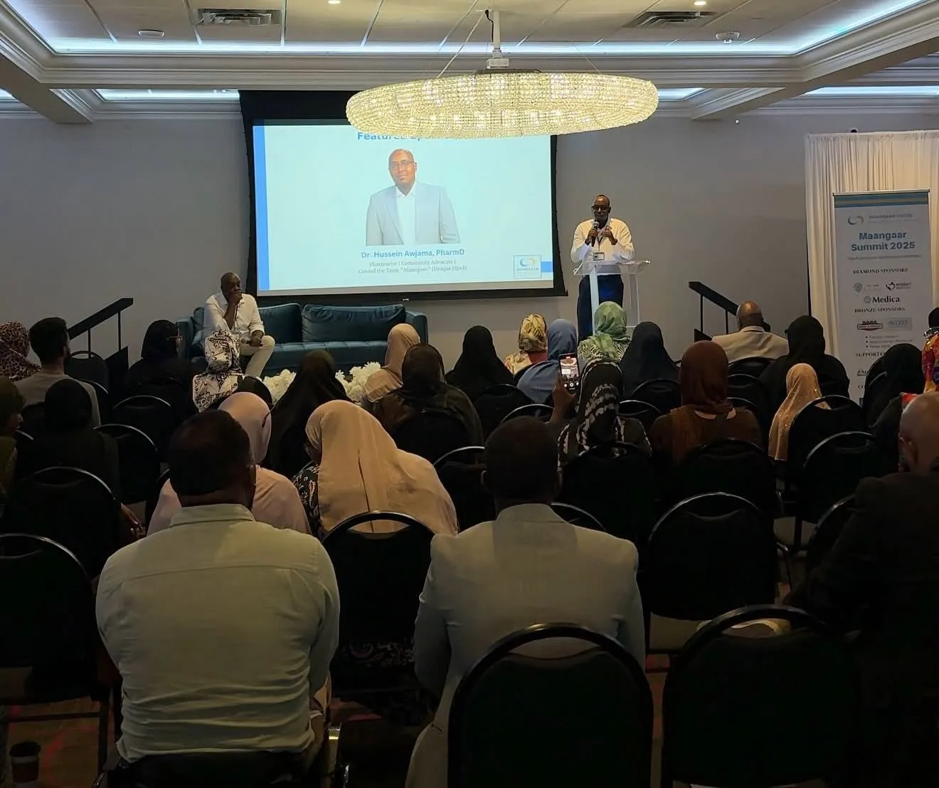 A full room faces the stage, where a speaker stands at the podium beneath a slide introducing Dr. Hussein Awjama, PharmD, pharmacist and community advocate. A Maangaar Summit 2025 sponsor banner stands at right.