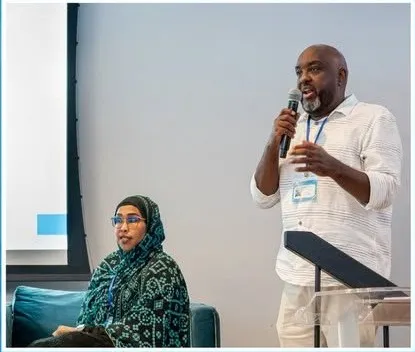 A speaker addresses the room with a microphone while a panelist listens.