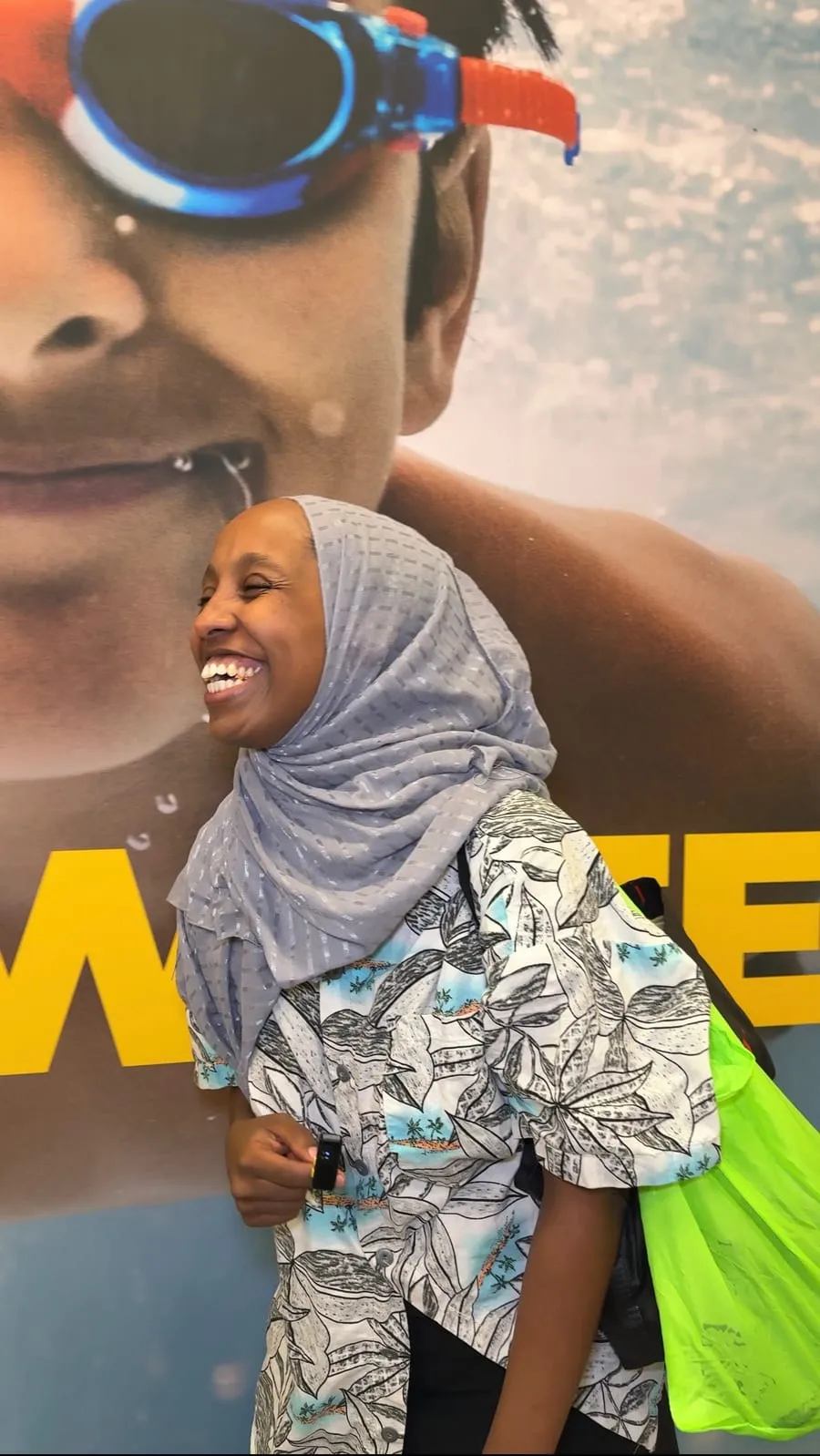 A participant in a headscarf laughs beside a swim school poster.